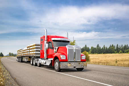 Bright red bonnet big rig semi truck with chrome accessories transporting fastened lumber cargo on flat bed semi trailer running on the narrow summer road stock photo